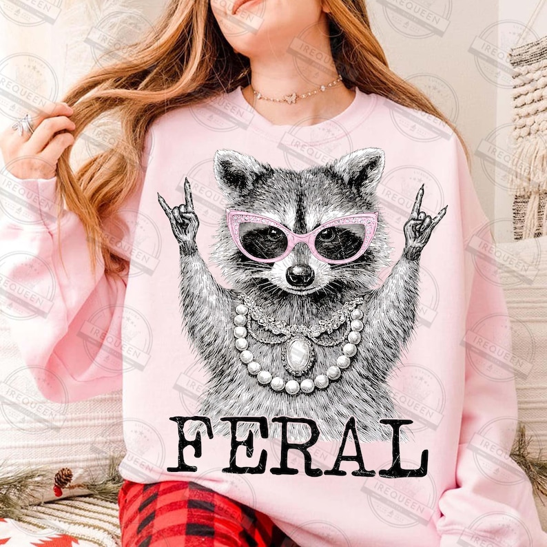 Puede incluir: Sudadera rosa claro con un mapache con gafas rosas y un collar de perlas. El mapache hace un gesto de rock and roll con la mano. La palabra "FERAL" est&aacute; impresa en letras negras.