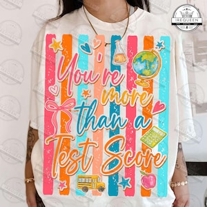 Może przedstawiać: Biała koszulka z kolorowym wzorem. Wz&oacute;r zawiera pionowe paski w kolorze r&oacute;żowym, pomarańczowym i niebieskim, z napisem "You're more than a Test Score". Znajdują się na niej r&oacute;wnież ilustracje autobusu szkolnego, jabłka i globusa.