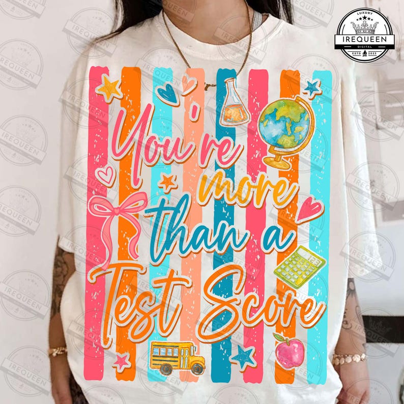 Może przedstawiać: Biała koszulka z kolorowym wzorem. Wz&oacute;r zawiera pionowe paski w kolorze r&oacute;żowym, pomarańczowym i niebieskim, z napisem "You're more than a Test Score". Znajdują się na niej r&oacute;wnież ilustracje autobusu szkolnego, jabłka i globusa.