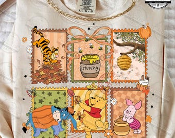 Puede incluir: Sudadera de color crema con un gráfico colorido de los personajes de Winnie the Pooh en un diseño de sello postal. El diseño incluye a Tigger, Pooh, Piglet, Eeyore, un tarro de miel y una colmena. La sudadera tiene el texto "COMFORT COLORS" en la parte superior.