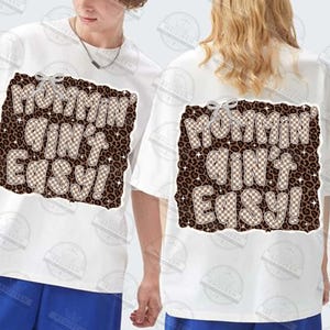 以下が含まれることがあります： 茶色のヒョウ柄と市松模様のデザインで「Mommin' Ain't Easy！」というフレーズがプリントされた白いTシャツ。半袖のシャツは、前面と背面にテキストが表示されています。
