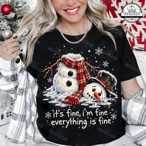 May include: A black t-shirt featuring a graphic of a melting snowman wearing a red and green plaid scarf and a black top hat. The design includes white snowflakes and the text 'it's fine, i'm fine, everything is fine' printed in white lettering.