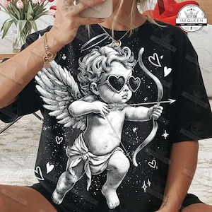 Op de afbeelding: Zwart T-shirt met een gedetailleerde illustratie van Cupido, de Romeinse god van de liefde, die een pijl afschiet. Cupido wordt afgebeeld met hartvormige zonnebrillen, vleugels en pijl en boog. Het ontwerp bevat harten en sterren.