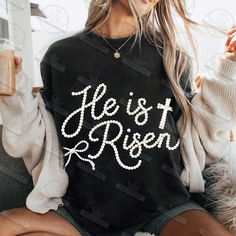 May include: Black crew neck t-shirt with the white text "He is Risen". The text is in a cursive font and is made of small white dots. The shirt is part of a casual wear collection.