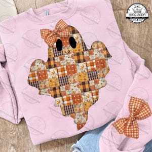 May include: A light pink sweatshirt featuring a patchwork ghost design with a bow. The ghost is made of various orange, brown, and cream-colored plaid and floral patterns. The bow on the ghost and the additional bow are orange and white checkered.