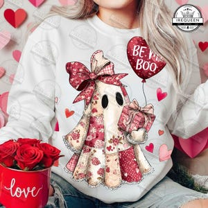 Könnte beinhalten: Ein weißes Sweatshirt mit einem Patchwork-Geist, der ein Geschenk und einen herzförmigen Ballon mit der Aufschrift "BE MINE BOO" hält. Der Geist hat eine große Schleife und ist von Herzen umgeben. Eine rote Tasse mit dem Wort "love" ist ebenfalls zu sehen.