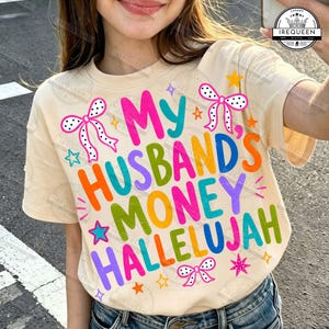 Pu&ograve; includere: Una t-shirt color crema con il testo colorato My Husband's Money Hallelujah in un carattere audace e giocoso. Il design &egrave; decorato con piccoli fiocchi a pois rosa e bianchi e varie forme di stelle gialle, rosa e blu.