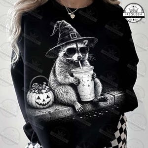 May include: A black long-sleeve sweatshirt featuring a graphic of a raccoon wearing a witch hat and sunglasses, holding a plastic cup with a straw. Next to the raccoon is a jack-o'-lantern pumpkin bucket filled with various candies.