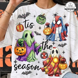 May include: A white t-shirt featuring a Halloween-themed graphic with the text 'tis the season'. The design includes three ghost figures dressed in superhero-inspired costumes, each holding a jack-o'-lantern, surrounded by bats and stars.