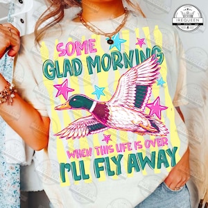 May include: A light beige t-shirt with a colorful graphic of a duck in flight, surrounded by stars. The text "SOME GLAD MORNING" and "WHEN THIS LIFE IS OVER I'LL FLY AWAY" are also displayed on the shirt.