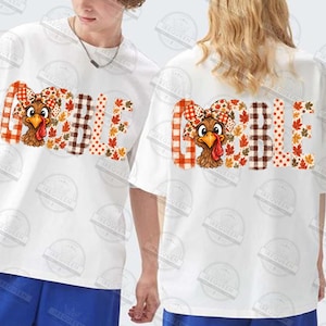 May include: A white short-sleeved t-shirt featuring the word GOBBLE printed in large letters. The letters are filled with a pattern of orange and brown plaid, polka dots, and autumn leaves. A cartoon turkey wearing a plaid bow is integrated.