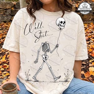 May include: A cream-coloured t-shirt featuring a black line art illustration of a headless skeleton walking while holding a skull-shaped balloon. The text "Well, Shit...." is printed in a cursive script above the skeleton, with small floral accents.