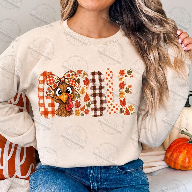 May include: A beige crewneck sweatshirt featuring the word GOBBLE printed across the chest. The letter O is replaced by a cartoon turkey wearing a plaid bow. The letters are decorated with orange plaid, brown gingham, and autumn leaf patterns.