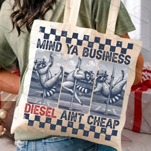 May include: A beige tote bag featuring a graphic of three raccoons pole dancing. The text "MIND YA BUSINESS" is above the raccoons, and "DIESEL AINT CHEAP" is below. The design includes a checkered pattern in navy blue and white.