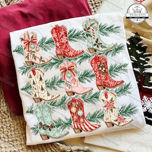 May include: A white t-shirt featuring a graphic print of eight cowboy boots decorated with Christmas themes, including snowflakes, pine trees, poinsettias, candy canes, and ornaments, all arranged among green pine needle branches.