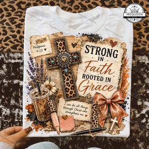 May include: A white t-shirt featuring a graphic print of a leopard print cross, a coffee cup, a bow, and text that reads Strong in Faith Rooted in Grace, Philippians 4:13, and I can do all things through Christ who strengthens me, with floral accents.