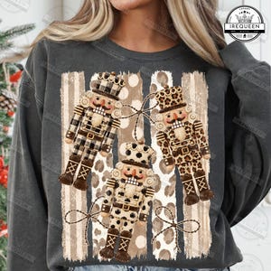 May include: A charcoal gray crewneck sweatshirt featuring a graphic print of three nutcrackers. The nutcrackers are decorated in black and white buffalo plaid, leopard print, and polka dot patterns, set against a background of vertical stripes.