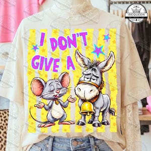 May include: A beige t-shirt featuring a graphic print of a grey mouse and a grey donkey wearing a gold bell. The text "I DON'T GIVE A" is printed in bright pink above the animals, surrounded by yellow vertical stripes and small purple stars.