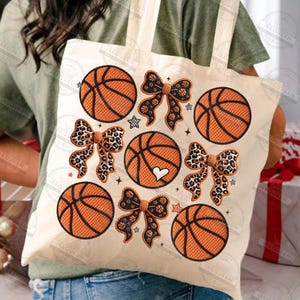 May include: A cream-colored tote bag featuring embroidered basketballs and bows. The basketballs are orange with black lines, and the bows have leopard print and black patterns. Small star and heart details are also included.