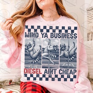 May include: A light pink sweatshirt with the text "MIND YA BUSINESS" above three raccoon illustrations. Below the illustrations, the text "DIESEL AINT CHEAP" is displayed. The design is framed by a checkered pattern.
