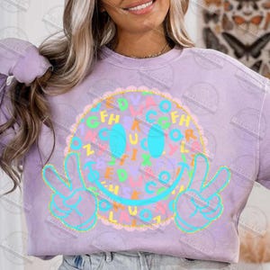 May include: A light purple sweatshirt featuring a large smiley face design. The smiley face is outlined in turquoise and has a peace sign gesture. The face is filled with colorful letters and a scalloped border.