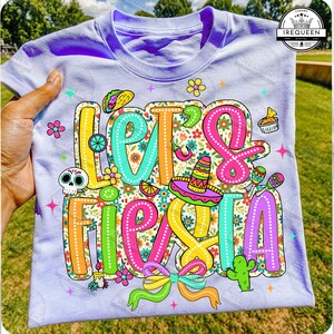 May include: A lilac t-shirt featuring the phrase "Let's Fiesta" in vibrant, outlined lettering. The design incorporates a sombrero, maracas, a cactus, and other celebratory graphics. The shirt is held by a hand.
