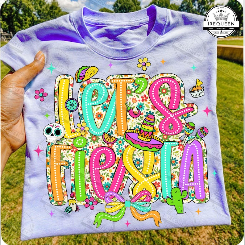 May include: A lilac t-shirt featuring the phrase "Let's Fiesta" in vibrant, outlined lettering. The design incorporates a sombrero, maracas, a cactus, and other celebratory graphics. The shirt is held by a hand.