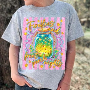 May include: A gray t-shirt featuring a colorful graphic design. The design includes a mason jar filled with fireflies, surrounded by flowers and stars, with the words "Fireflies and front porch nights" in a whimsical font.