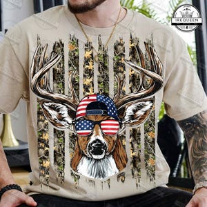May include: Beige t-shirt featuring a deer head wearing a cap and sunglasses with an American flag design. The deer has large antlers and is set against a camouflage and flag background. The shirt has a crew neck.