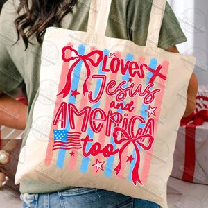 Puede incluir: Bolso tote crema con el texto "Loves Jesus and America too" en rojo y azul. El dise&ntilde;o incluye una bandera estadounidense, estrellas y lazos rojos, con rayas verticales rojas y azules.