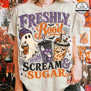 Op de afbeelding: Een crèmekleurig t-shirt met een afbeelding van een geest met een paarse muts die een pompoendrankje vasthoudt, naast twee ijskoffiebekers. De tekst luidt "FRESHLY BOO'd with SCREAM & SUGAR" in paarse en oranje letters.