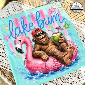 May include: A light blue t-shirt with the words "lake bum" in blue script. The shirt features a cartoon Bigfoot wearing sunglasses and a red cap, lounging on a pink flamingo float and holding a coconut drink. Pink and yellow stars decorate the background.