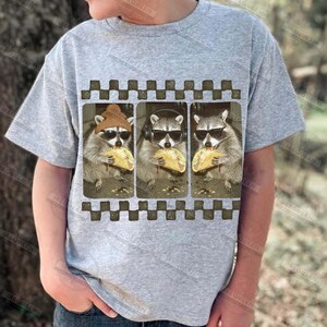 May include: A light grey t-shirt with a graphic of three raccoons wearing sunglasses and eating tacos. The raccoons are in a framed design with a checkered border. The text "IREQUEEN" is printed on the shirt.