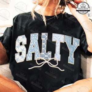 Puede incluir: Camiseta negra con la palabra "SALTY" en letras grandes de color azul claro. Las letras están decoradas con patrones de conchas marinas y estrellas de mar. Un lazo de perlas está debajo de la palabra. Diseño casual de playa.