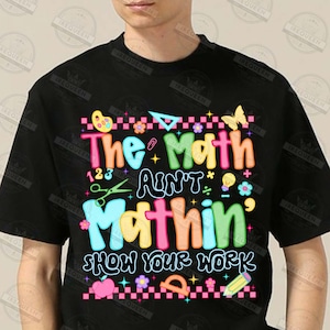 Puede incluir: Camiseta negra con la frase "The Math Ain't Mathin' Show Your Work" en letras coloridas y divertidas. El dise&ntilde;o incluye s&iacute;mbolos matem&aacute;ticos, una mariposa, tijeras y un l&aacute;piz, ideal para profesores o estudiantes.