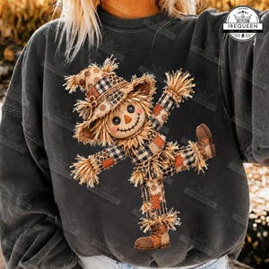 May include: A charcoal gray sweatshirt featuring a graphic of a scarecrow with a stitched smile, button eyes, and a triangular nose. The scarecrow wears a patchwork outfit of plaid, leopard print, and polka dot patterns with straw accents.