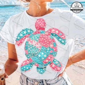 May include: White t-shirt featuring a colorful patchwork turtle design. The turtle is composed of pink and turquoise fabric with floral and star patterns. The design is centered on the shirt, creating a vibrant and eye-catching look.