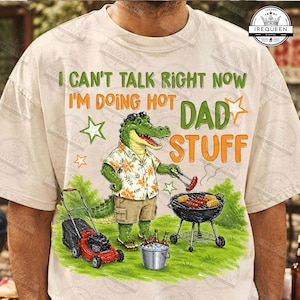 May include: A beige t-shirt featuring a green alligator wearing a floral shirt and khaki shorts, standing next to a red lawnmower, a charcoal grill with sausages, and a bucket of beer. The text reads: I CAN'T TALK RIGHT NOW I'M DOING HOT DAD STUFF.