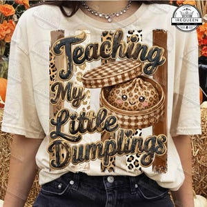May include: A cream-colored t-shirt featuring a graphic print of a cute dumpling with a leopard print pattern inside a bamboo steamer basket. The text reads "Teaching My Little Dumplings" in a bold, dark font over vertical leopard print stripes.