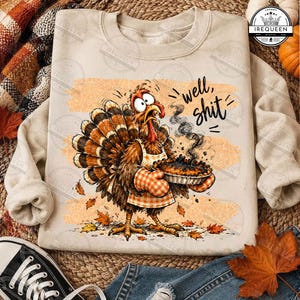 May include: A beige crewneck sweatshirt featuring a graphic of a cartoon turkey wearing an apron and checkered oven mitts, holding a steaming burnt pie. The text "well, shit" is printed in black script above the turkey, surrounded by orange leaves.
