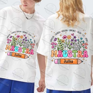 May include: White t-shirt with a colorful graphic of the word "MAESTRA" surrounded by flowers and butterflies. The graphic also includes a pencil with the name "Jullia" written on it. The text around the word "MAESTRA" reads "compasivia, cari&ntilde;osa, dedicada, confiable, c&aacute;lida, leal, amable".