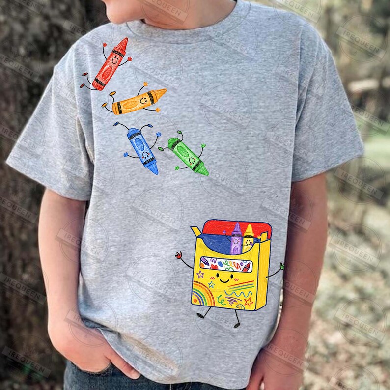 May include: A heather grey t-shirt featuring a graphic print of a yellow crayon box with a smiling face and arms and legs. Above the box, four individual colourful crayons in red, orange, blue, and green are illustrated with faces and limbs.