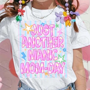 May include: White t-shirt with the text "JUST ANOTHER MANIC MOM-DAY" in pink, outlined in white. The design includes stars, lightning bolts, and flowers in various colors. The background has vertical blue and white stripes.