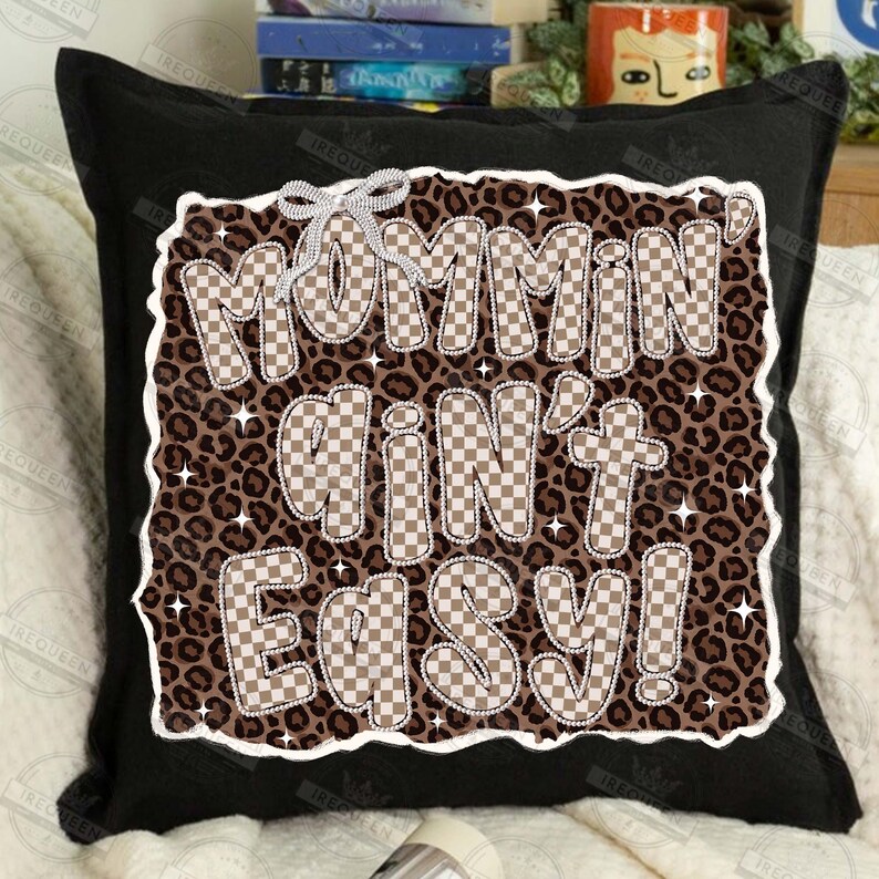 以下が含まれることがあります： 黒い正方形のクッション。テキスト「Mommin' Ain't Easy！」は市松模様で、ヒョウ柄の背景に配置されています。クッションには白い縁取りと装飾用のリボンが付いています。