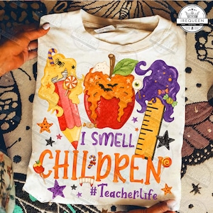 May include: A white t-shirt featuring a graphic of a red apple, a yellow pencil, and a wooden ruler, all topped with colorful melting slime. The text reads I SMELL CHILDREN and #Teacherlife, surrounded by small stars, candies, and a spider.