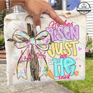 May include: A cream-colored sweatshirt featuring a cross with a pink and yellow bow, Easter eggs, and the text "He has Risen Just as He said" and "Luke 24:6". The design includes cartoon bunnies and a carrot.