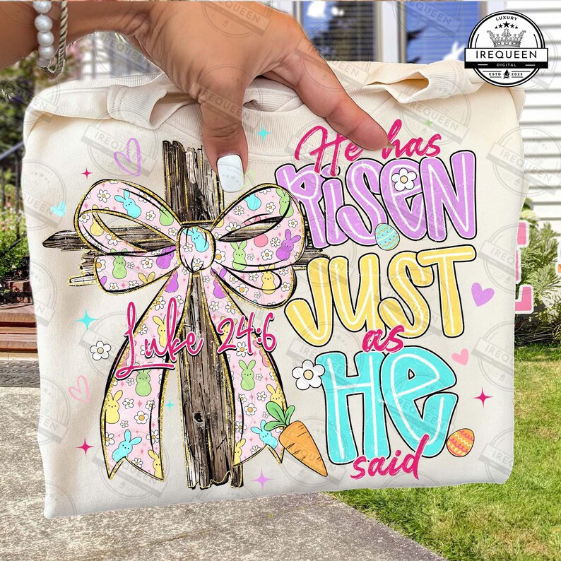 May include: A cream-colored sweatshirt featuring a cross with a pink and yellow bow, Easter eggs, and the text "He has Risen Just as He said" and "Luke 24:6". The design includes cartoon bunnies and a carrot.