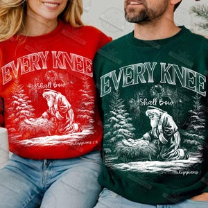 May include: Red and green Christmas sweatshirts with the text "EVERY KNEE Shall Bow" in white. Each sweatshirt features a graphic of Santa kneeling before a manger scene, surrounded by pine trees. The text "Philippians 2:10" is also visible.