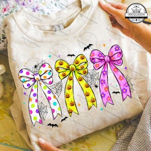 May include: A beige crewneck sweatshirt featuring a graphic print of three large bows. The left bow is white with colorful polka dots, the center bow is yellow with orange jack-o'-lantern faces, and the right bow is pink with purple flowers.