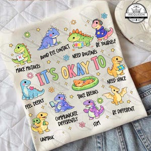 May include: A white t-shirt with cartoon dinosaurs and phrases such as "It's Okay To" and "Take Breaks." The design incorporates flowers and stars, offering a cheerful and encouraging aesthetic.